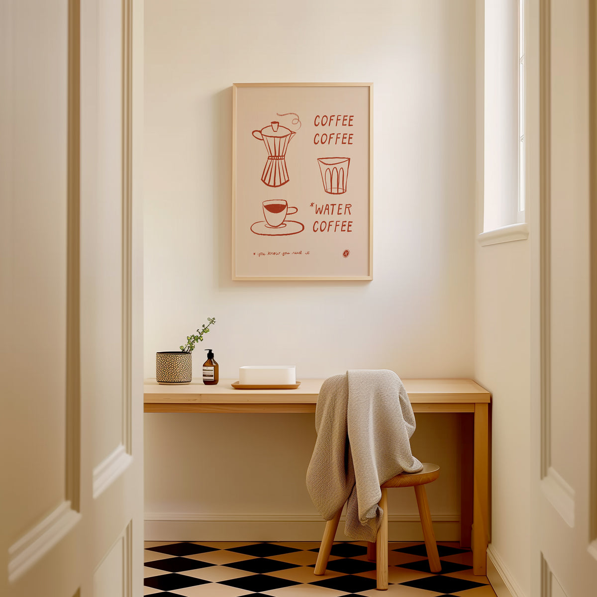 A framed minimalist illustrated moka pot, espresso cup, and water glass line art print in rust red on cream with text spelling out COFFEE, COFFEE, WATER, COFFEE, hangs above a wooden desk in a bright, modern room with a checkered floor and a chair draped with a gray blanket.