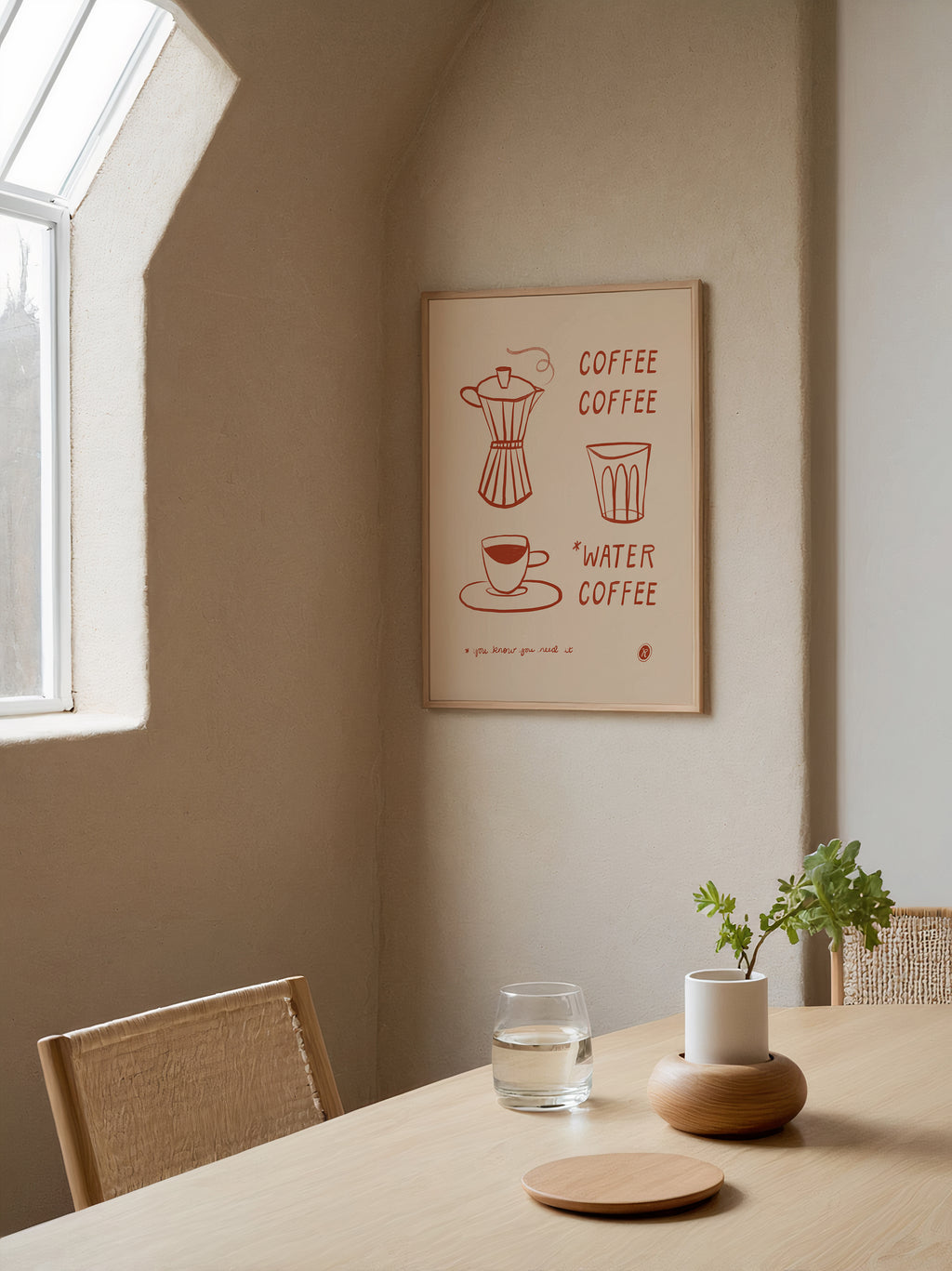 A framed minimalist coffee-themed wall art print hangs on a beige wall in a sunlit dining room. The print features simple red line drawings of a moka pot, a cup, and the words 'COFFEE' and 'WATER COFFEE'.