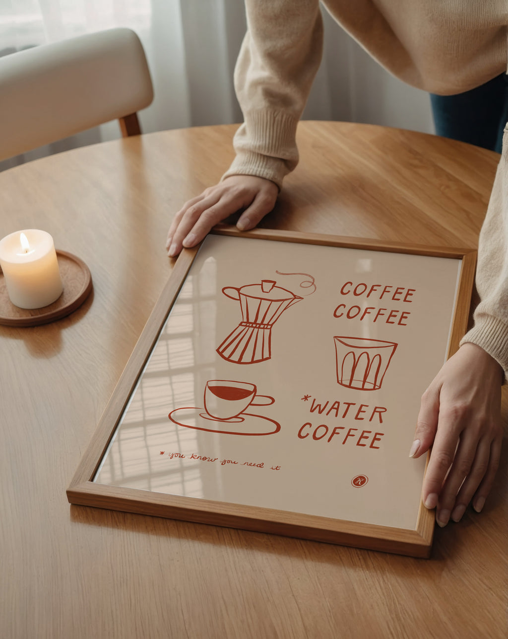 A framed coffee-themed art print with minimalist red line drawings and text, held on a wooden table by a person wearing a beige sweater. A lit candle sits nearby in a softly lit interior setting.