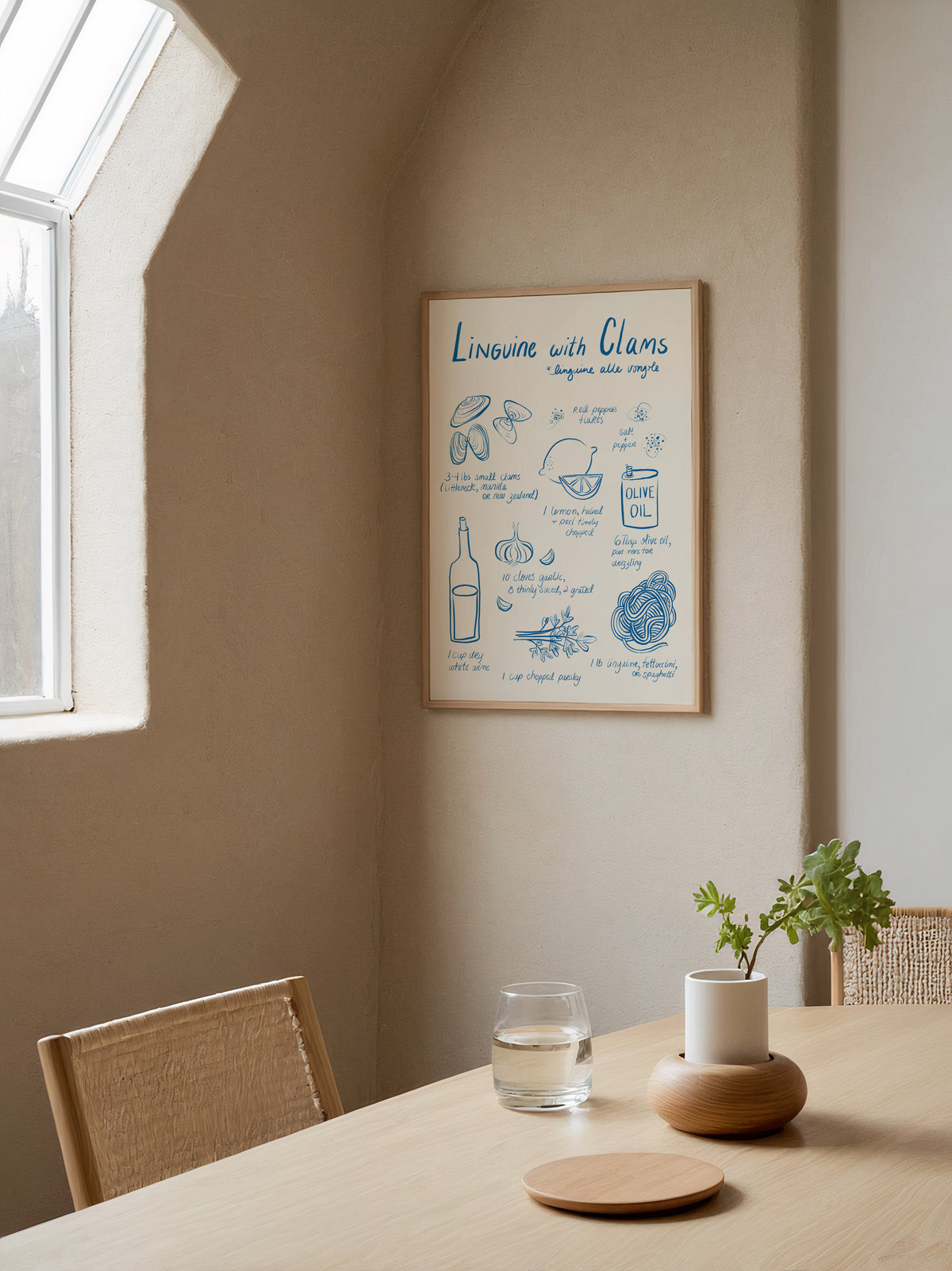 A framed recipe-style print titled 'Linguine with Clams' hangs on a beige wall in a sunlit dining corner. The print has blue illustrated notes and text on a cream background.