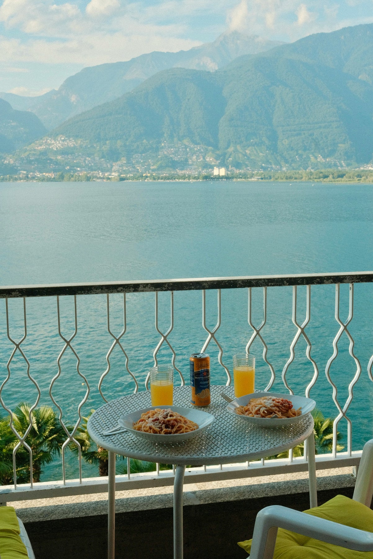 A scenic view of a large blue lake and green mountains from a balcony, with a small round table in the foreground holding two plates of pasta, two glasses of orange juice, and a can of beverage.
